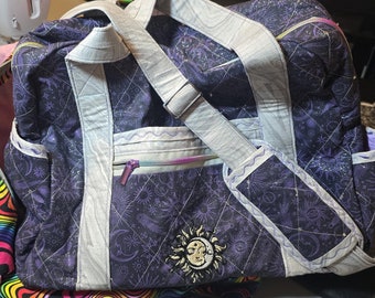 May include: A purple and white quilted duffel bag with a celestial print and a sun and moon design. The bag has a zipper closure and a shoulder strap.