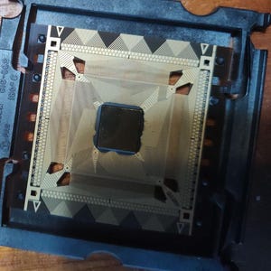 Puede incluir: Primer plano de un procesador de computadora, con una unidad central de procesamiento (CPU) dorada y circuitos intrincados. La CPU está dentro de un marco de plástico negro, mostrando su diseño complejo y componentes tecnológicos.