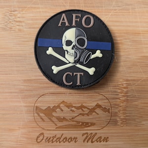 Può includere: Una toppa nera e circolare con un teschio e una maschera antigas, con ossa incrociate sotto. Il testo "AFO CT" è sopra e sotto il teschio. Una linea blu attraversa il teschio. La toppa è su una superficie di legno.