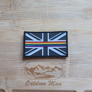 Può includere: Una toppa rettangolare con il design della bandiera Union Jack e una striscia arcobaleno. La toppa ha un bordo nero ed è posta su una superficie di legno. Il testo "Outdoor Man" è visibile sulla superficie di legno.