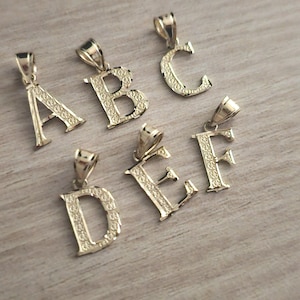 May include: Six gold-tone pendants, each featuring a different letter of the alphabet, A through F. The pendants are intricately detailed with a decorative pattern.