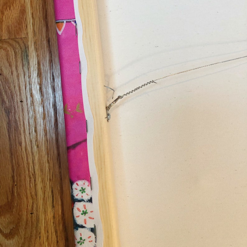 May include: The back of a canvas painting with a wooden frame. A wire is attached to the frame with a hook for hanging.