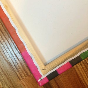 May include: Close-up of a corner of a canvas print with a wooden frame. The canvas is stretched tightly over the frame and secured with staples. The canvas is printed with a colorful abstract design.
