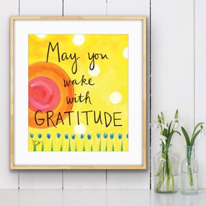 May include: A framed print with a yellow background and a pink and orange sun. The text reads "May you wake with GRATITUDE".