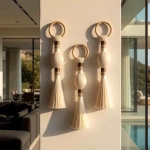 Pode incluir: Três penduradores de parede decorativos com uma estética boêmia natural. Cada um apresenta um topo circular, uma esfera central e uma longa borla creme. Estão montados numa parede clara, com um interior moderno e vista para o mar ao fundo.
