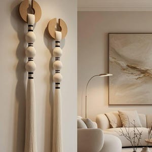 Op de afbeelding: Twee wanddecoraties met licht houten cirkelvormige bovenkanten met een centrale gleuf. Elk stuk heeft een verticale opstelling van drie gebroken witte stoffen bollen gescheiden door dunne zwarte banden, eindigend in een lange, rechte gebroken witte kwast.