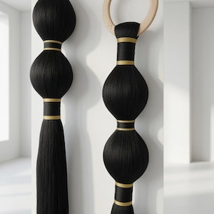 May include: Two vertical wall hangings made of black thread wrapped around spherical forms, accented with horizontal gold bands. The right piece features a circular light-coloured wooden ring at the top, while both terminate in long, straight black tassels.