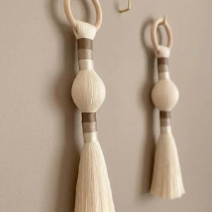 May include: Two decorative wall hangings featuring a light-colored wooden ring at the top, connected to a central bulbous shape wrapped in cream and brown thread, ending in a long, straight, cream-colored fringe tassel hanging vertically.