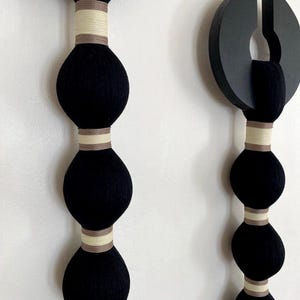May include: Two vertical wall hangings featuring a series of black yarn-wrapped spheres separated by bands of light beige and tan woven fabric. Each piece is topped with a black circular wooden element with a cutout, creating a structured design.