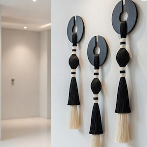 May include: Three contemporary wall decorations. Each has a black circular top, a black sphere, and a cream-coloured tassel. The design includes white and black stripes. The wall hangings are mounted on a white wall.