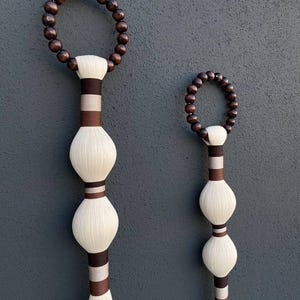 May include: Two decorative wall hangings. Each features a dark brown beaded circle at the top, connected to a white, bulbous body with brown and tan accents. The bottom of each piece has a flowing, white tassel.