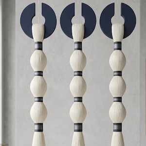 Può includere: Tre decorazioni da parete verticali sospese con cime circolari in legno blu scuro. Ogni pezzo è composto da una serie di segmenti in fibra color crema, testurizzati e sferici, separati da sottili fasce orizzontali grigio scuro in una struttura a colonna.
