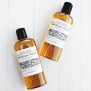 May include: Two amber glass bottles of body wash with black lids and white labels. The labels read "body wash Wildflower Honey botanical formula 8 oz net wt".
