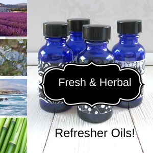 May include: Four small blue glass bottles with black lids and labels. The labels have a black and white design and the text "Fresh & Herbal Refresher Oils!" The bottles are arranged in a row on a white wooden surface.