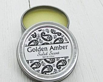 May include: A small silver tin with a black and white paisley pattern label. The label reads "Golden Amber Solid Scent Net wt 1/2oz".