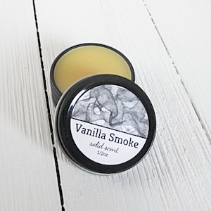 May include: A round black metal tin containing a pale yellow solid scent. The lid features a white label with a black and white abstract smoke pattern, the text Vanilla Smoke, the words solid scent, and the measurement 1/2oz.