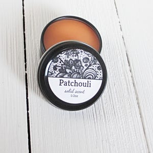 May include: A round black metal tin containing amber-colored solid scent. The lid features a white label with a black lace-like floral pattern, the text Patchouli, solid scent, and 1/2oz printed in black ink on the front of the container.