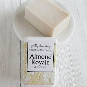 May include: A bar of Almond Royale handcrafted soap, 4.5 oz, rests on a white soap dish. The soap is light beige and rectangular. The soap packaging features the text "gently cleansing" and "Almond Royale".