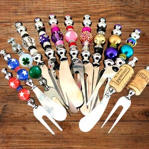 May include: A collection of decorative cheese spreaders and forks with ornate, beaded handles. The handles feature various designs, including red, white, and blue star-studded balls, green spheres, and sparkling beads. The utensils are arranged on a wooden surface.