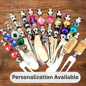 May include: A collection of decorative cheese spreaders and forks with various beaded handles in multiple colors. Some handles feature corks. The image includes the text "Personalization Available".
