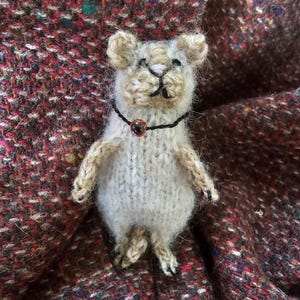 May include: A small, hand-knitted figurine of a mouse crafted from fuzzy, off-white and light brown yarn. The mouse features a black thread collar with a single, small, round, reddish-brown bead at the center, and has dark stitched facial details.