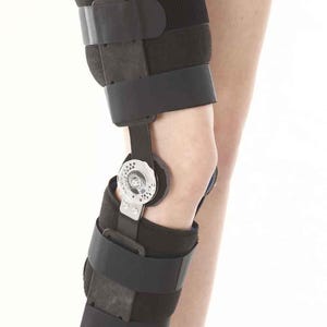 May include: A black hinged knee brace featuring adjustable fabric straps and a central metal dial mechanism. The support structure consists of padded sections above and below the knee joint, connected by rigid side bars with a circular hinge.