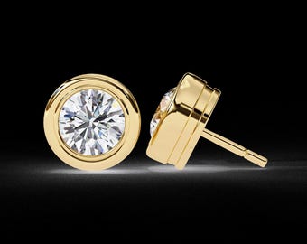 May include: Two gold bezel-set stud earrings with a brilliant-cut diamond. The earrings are round with a bright, reflective surface. The post and backing are visible on one earring. The background is black.