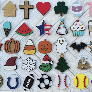 May include: A collection of 36 wooden shapes painted in various colors and designs. The shapes include a house, a cross, a heart, a shamrock, an Easter egg, a bunny, a flower, a sun, a popsicle, an ice cream cone, a watermelon, an American flag, a Christmas tree, a cupcake, a coffee cup, a camper, a sunflower, a maple leaf, a pumpkin, a candy corn, a jack-o-lantern, a ghost, a bat, a spider web, a turkey, a Christmas ornament, a snowman, a Santa hat, a snowflake, the Grinch, a Christmas tree, a couple, a wreath, a horseshoe, a football, a soccer ball, a baseball, a softball, a volleyball, and a basketball.