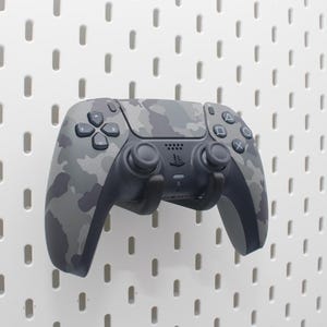 May include: A grey camouflage-patterned games controller with a black base is mounted on a white pegboard. The controller has various buttons and joysticks, and the PlayStation logo is visible.