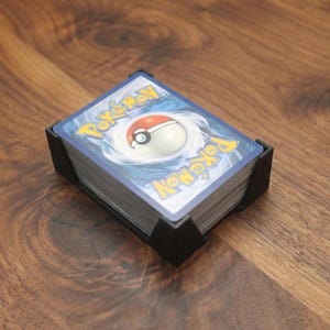 May include: A stack of Pokemon trading cards in a black card holder. The cards have a blue and white design with the Pokemon logo. The holder is on a wooden surface.
