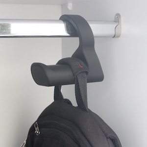 May include: A black backpack hanging from a black hook attached to a metal rod inside a white cabinet. The hook is designed to hold the backpack's straps, keeping it off the floor. The hook is made of a durable plastic material.