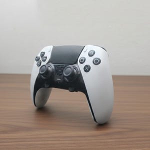 May include: A white and black wireless game controller with a central touchpad and button layout. The controller features a sleek design with a black center section and white handles. The buttons include a directional pad, action buttons, and analog sticks.