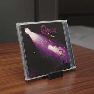 Może przedstawiać: Album CD zespołu Queen w przezroczystym plastikowym etui, prezentowany na drewnianej powierzchni. Okładka albumu przedstawia reflektor sceniczny z nazwą zespołu w kolorze różowym. Płyta CD jest podparta małym czarnym stojakiem.