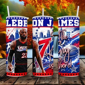 May include: A cylindrical stainless steel tumbler featuring a graphic of LeBron James in a black Philadelphia 76ers jersey, the text "LEBRON JAMES" and "The King is here!", a basketball, city skyline, and red, white, and blue star accents with a straw.