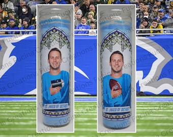 Könnte beinhalten: Zwei weiße Gebetskerzen mit blau-weißem Design. Eine Kerze zeigt einen Mann, der ein Buch mit dem Wort "EMINEM" darauf hält. Die andere Kerze zeigt denselben Mann, der einen Football hält. Beide Kerzen tragen den Text "ST. JARED OF DETROIT".