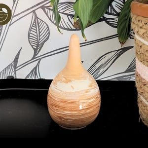 Peut inclure: Un vase en céramique avec une base arrondie et un long col fin. Le vase présente un motif marbré dans les tons de marron et de crème. Le vase est posé sur une surface noire, avec un panier tressé et une plante feuillue en arrière-plan.