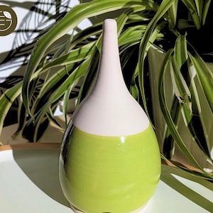 Peut inclure: Un vase en céramique fait à la main avec une base verte bombée, une section médiane blanche et un col rose pâle. Le vase a une finition brillante et une forme allongée unique. Des plantes vertes sont en arrière-plan.