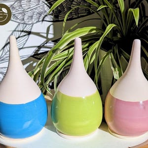 Peut inclure: Trois vases en céramique en forme de goutte d'eau. Chaque vase a un sommet blanc et une base colorée en bleu, vert et rose. Les vases sont exposés sur une surface en bois de couleur claire.