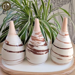 Peut inclure: Trois vases en céramique avec une base crème et des motifs de coups de pinceau bruns. Les vases ont un col étroit rose clair et sont disposés sur un plateau blanc avec un bord en bois. L'arrière-plan présente une plante et du papier peint à motifs.
