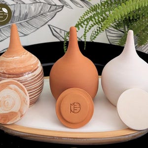 Peut inclure: Trois vases en céramique de couleurs terre cuite et blanche, avec des dessous de verre ronds assortis. Les vases ont une forme bulbeuse avec un long col fin. Un vase présente un motif rayé. Les dessous de verre et un vase portent les initiales "ME".