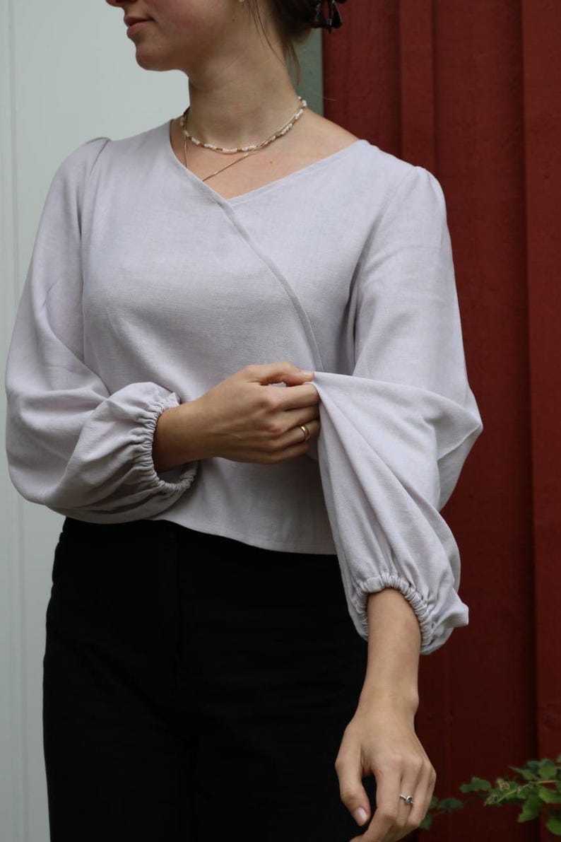 May include: A light purple, long-sleeved blouse with a V-neck and gathered sleeves. The blouse is made of a soft, lightweight fabric.