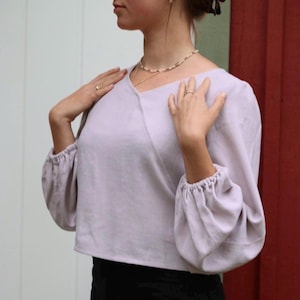 May include: A light purple, cropped blouse with long sleeves and a gathered detail at the wrists. The blouse has a V-neckline and a relaxed fit.