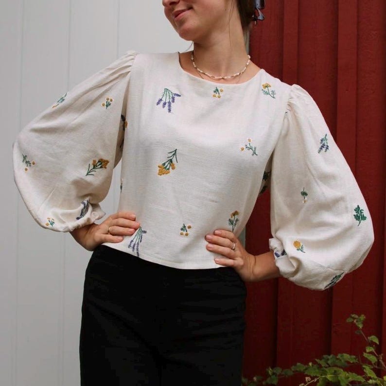 May include: A white blouse with a floral embroidery design. The blouse has a round neckline and long, puffy sleeves.