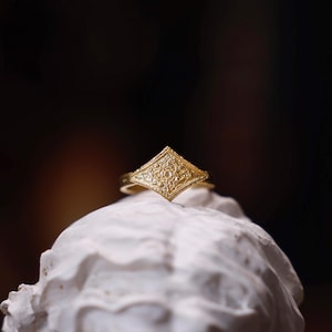 Pode incluir: Um anel de ouro com uma configuração em forma de diamante que apresenta um design floral intrincado.
