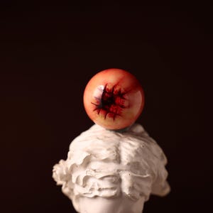 Puede incluir: Una escultura de busto blanco con un objeto rojo en forma de esfera en la parte superior. La esfera tiene un centro oscuro y texturizado y un acabado brillante. El fondo es marrón oscuro.