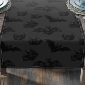 Puede incluir: Un camino de mesa negro con un patrón repetido de murciélagos. Los murciélagos son de color gris oscuro y están dispuestos en un patrón simétrico.