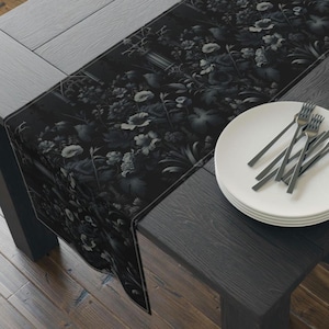 Könnte beinhalten: Ein schwarzer Tischläufer mit floralem Muster und detailliertem botanischem Design mit Blumen und Blättern. Der Läufer liegt auf einem dunklen Holztisch, mit weißen Tellern und Gabeln auf der rechten Seite. Die Maße sind nicht sichtbar.