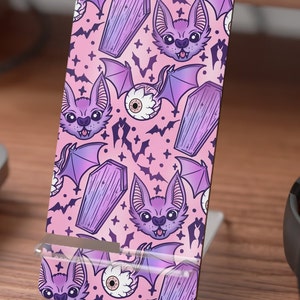 Può includere: Una custodia per telefono rosa con un motivo ripetitivo di pipistrelli viola, bare e occhi. I pipistrelli hanno ali viola e sorridono. Le bare sono viola e hanno una texture di legno. Gli occhi sono bianchi con pupille nere.