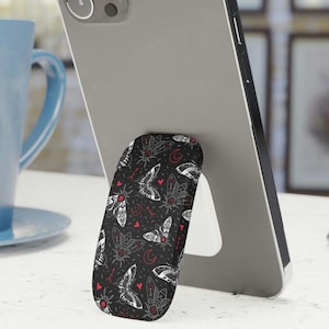 May include: A black phone stand with a pattern of white moths and red hearts. The stand is designed to prop up a phone.