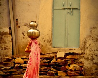 Puede incluir: Una mujer con un sari de rayas rosa y blancas lleva dos grandes vasijas de agua redondas y plateadas en la cabeza. Camina junto a una pared de piedra y una puerta azul con una reja de metal.